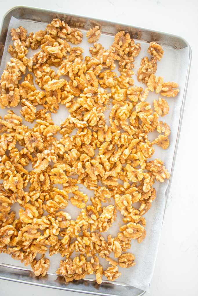 raw walnuts coated with hot honey on a baking sheet on a white marble countertop.