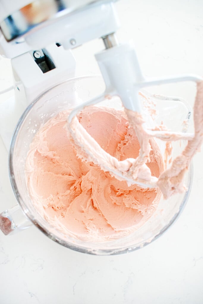 pink buttercream frosting in a glass mixing bowl on a white marble counter.