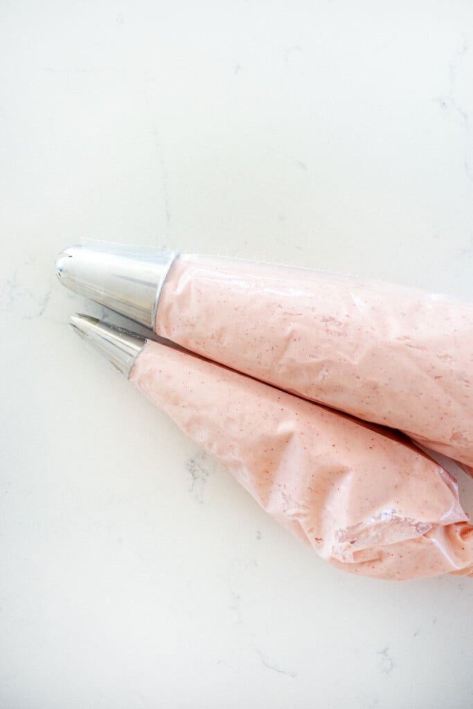 pink frosting in piping bags with decorating tips on a white marble counter.