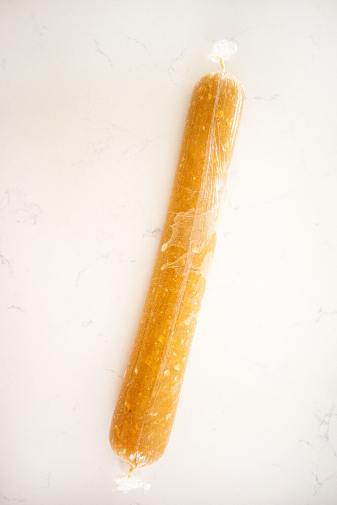 peanut butter oatmeal cookie dough rolled into a log and wrapped in plastic wrap on a white marble counter.