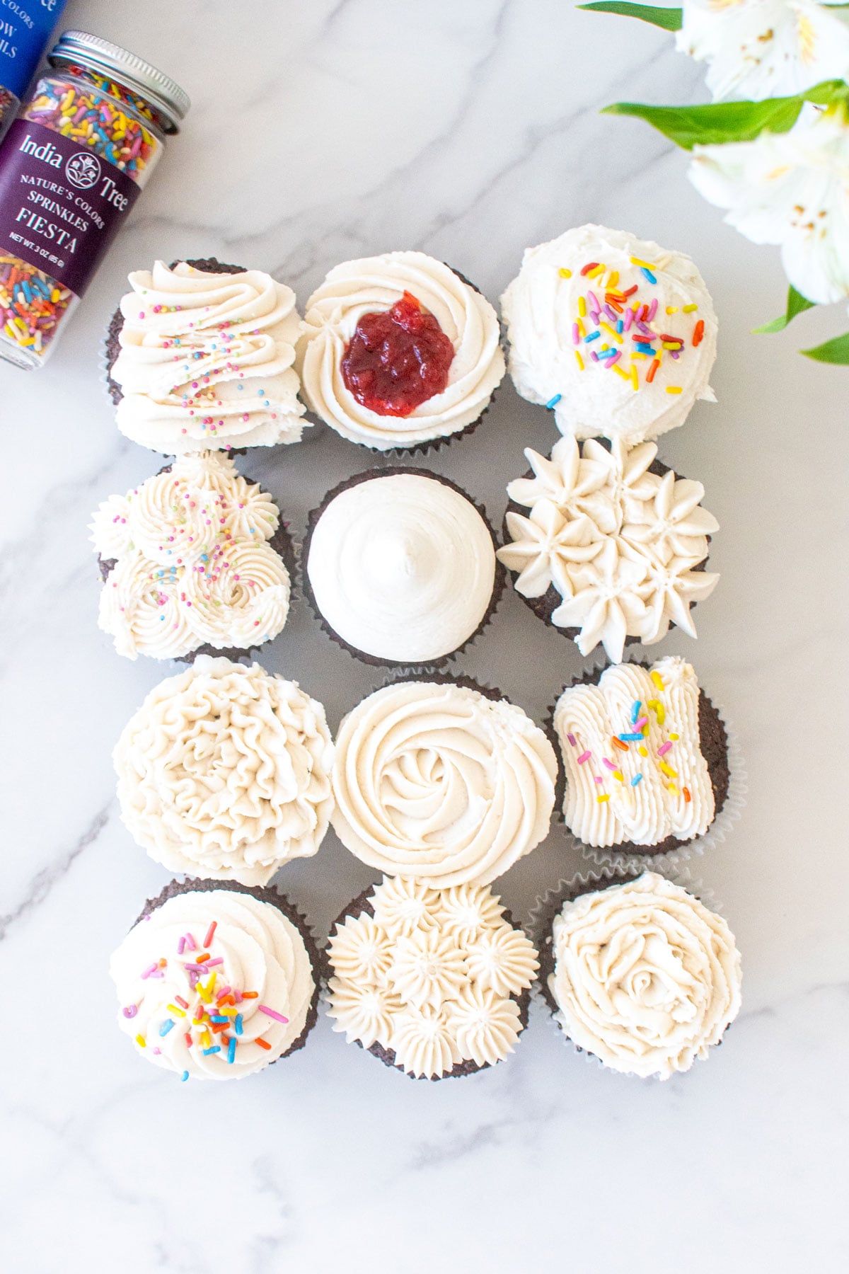 vegan chocolate cupcakes frosted 12 different ways on a white marble table with flowers and sprinkles.