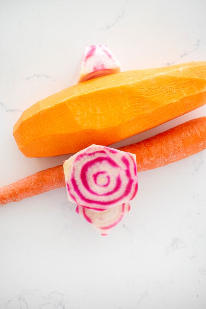 peeled sweet potato, beets, and carrots on a white countertop.