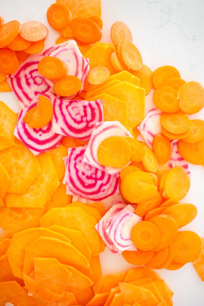 thinly sliced sweet potatoes, beets, and carrots on a white countertop.