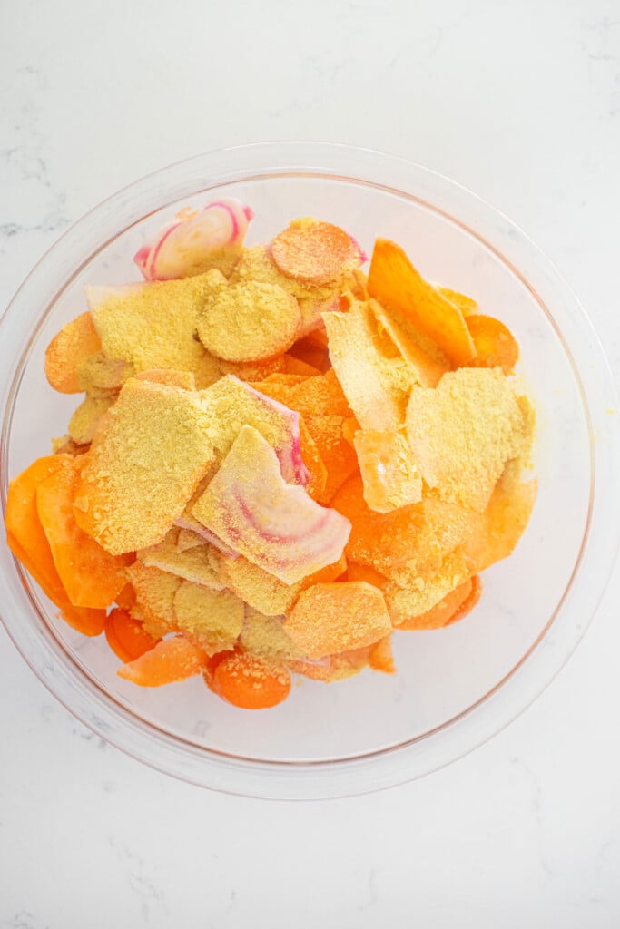 sliced sweet potato, beats, and carrots in a bowl with coconut oil and nutritional yeast on a whtie countertop.