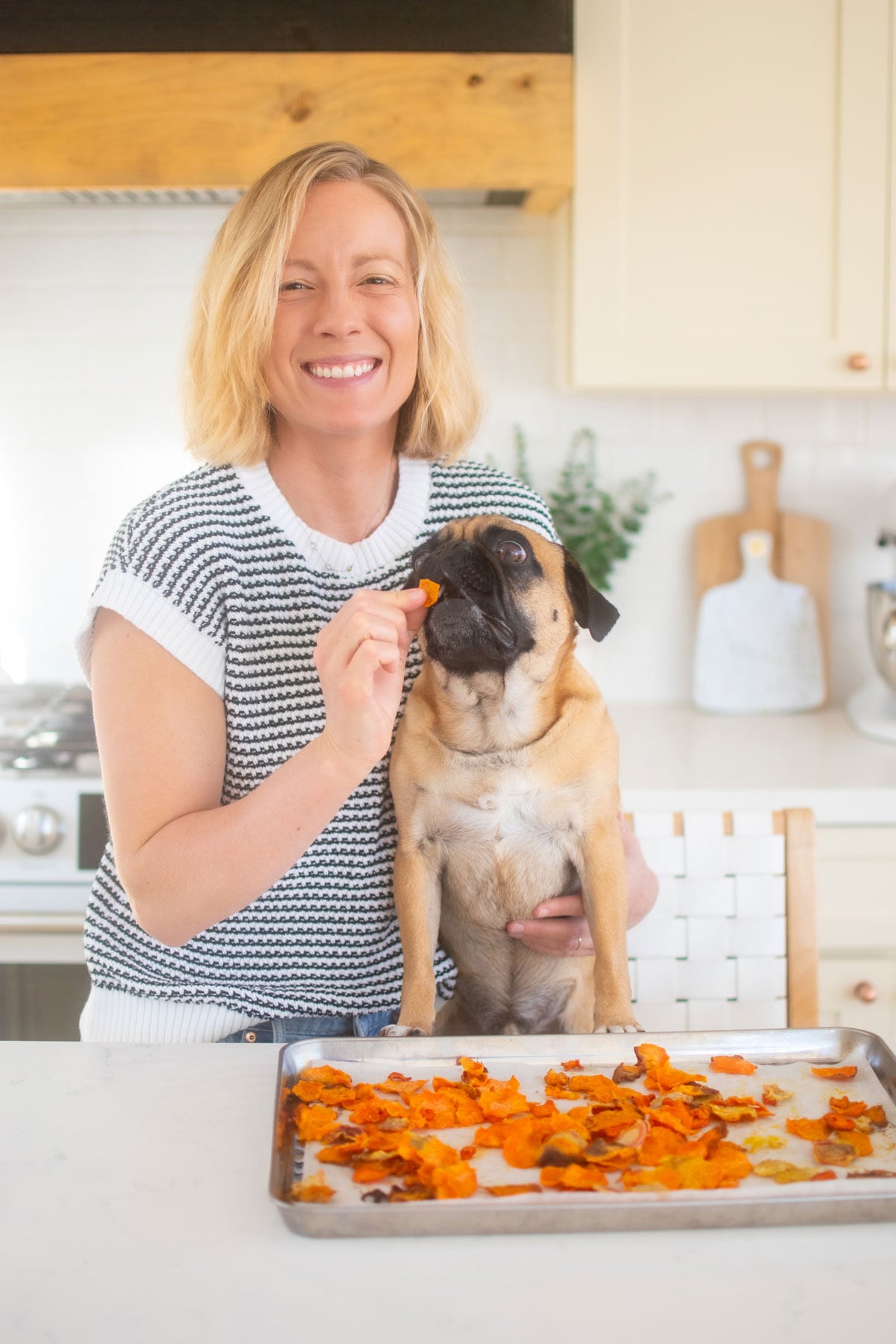 woman feeding pug veggie dog treats at the kitchen counter.