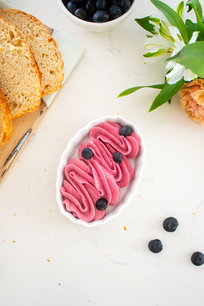 small dish with blueberry butter spread piped into a swirl sitting on a marble counter with bread and fresh flowers.