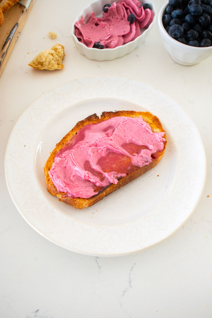 toasted sourdough with blueberry butter spread on top on a plate on a marble counter with a bowl of blueberries.