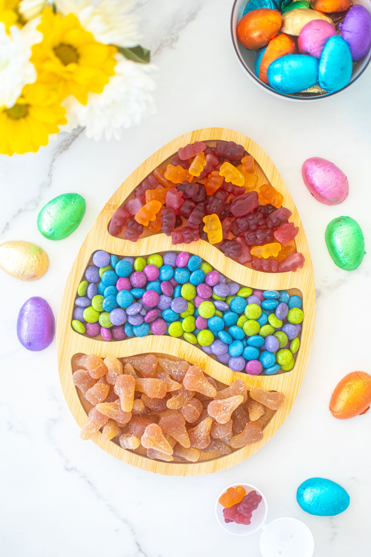 easter egg wood serving tray filled with easter candy on a white marble counter with fresh flowers.