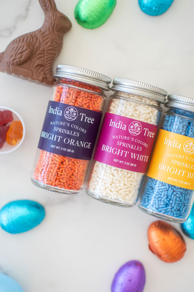 india tree nature's colors orange, white and blue sprinkle jars on a white marble counter with chocolate bunny and foiled wrapped chocolate eggs.
