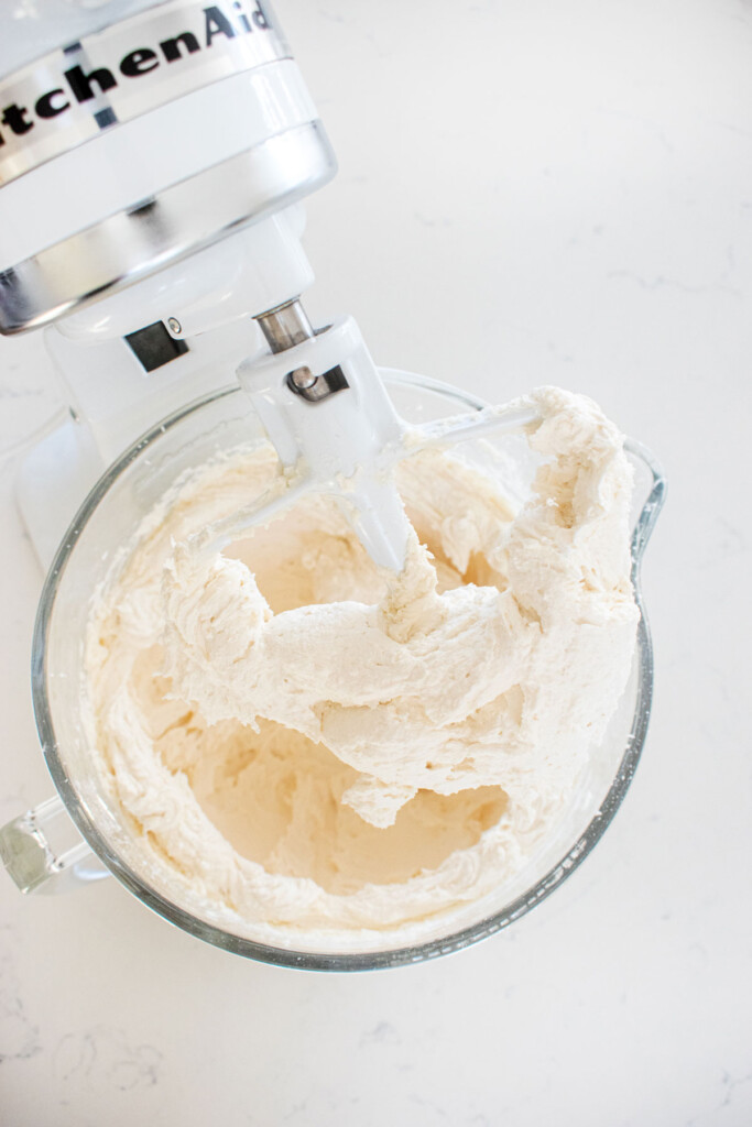 buttercream frosting in a glass mixing bowl on a white marble counter.
