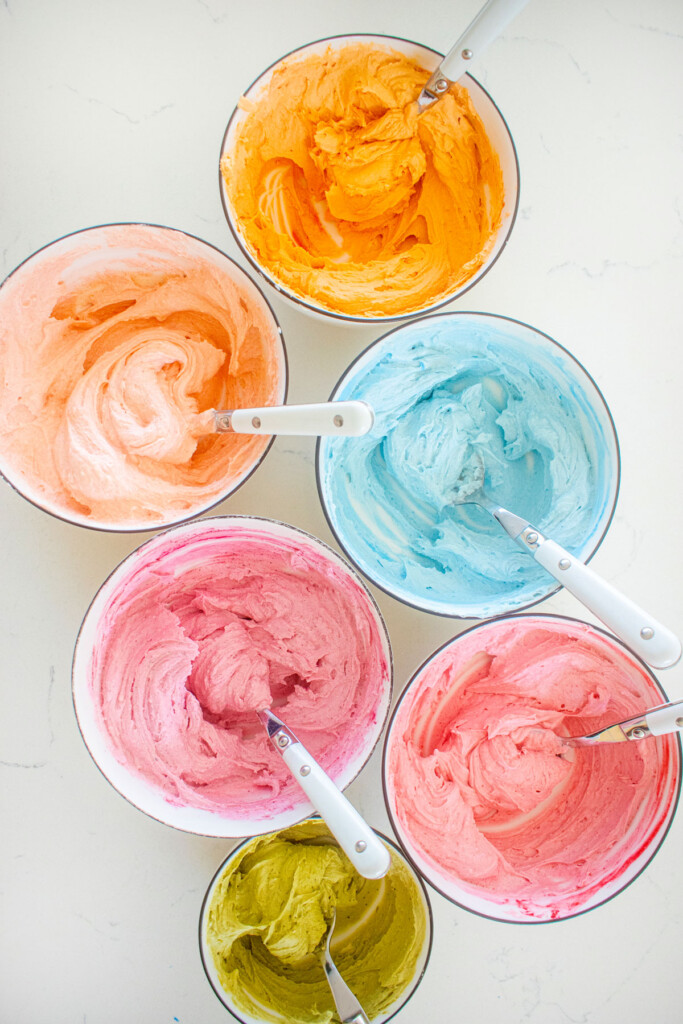 6 bowls with different frosting colors, blue, orange, pink, raspberry, and coral on a white marble counter.