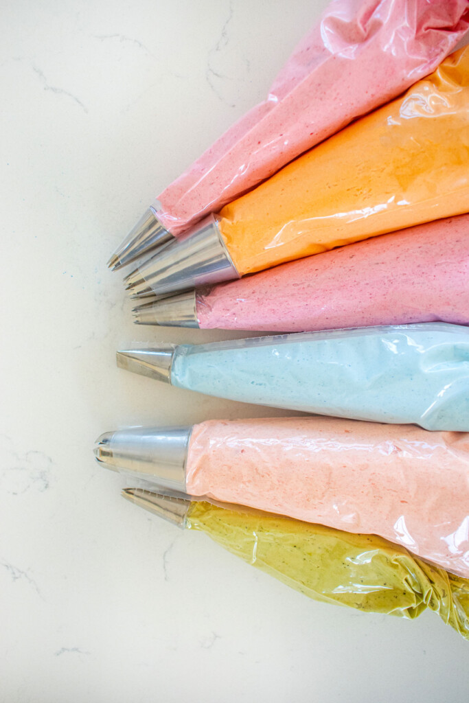 piping bags of frosting colors blue, orange, pink, raspberry, and coral on a white marble counter.
