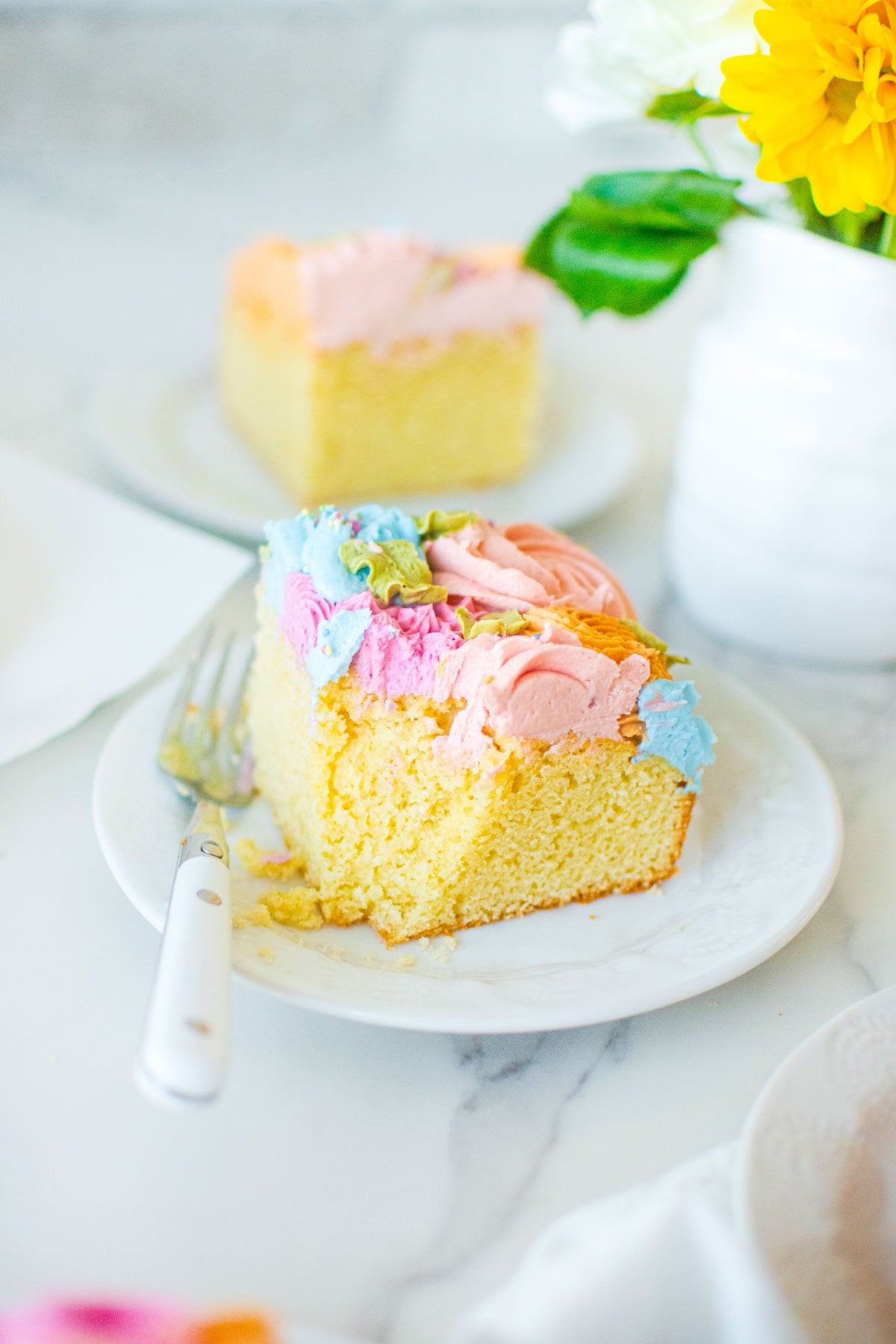 slice of pastel colored flower sheet cake with a bite taken out on a plate on a white marble counter.
