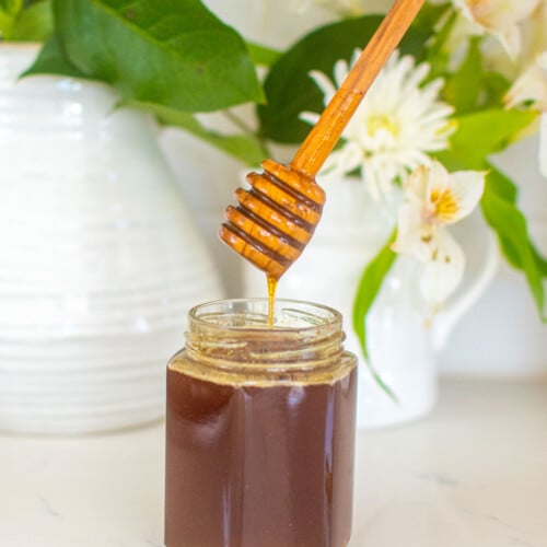 homemade organic hot honey in a glass honey jar with a honey dipstick on a marble counter with fresh flowers.
