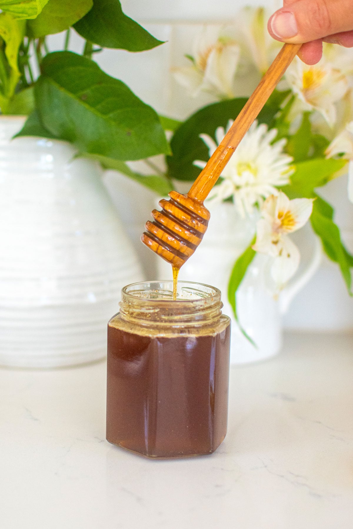 homemade organic hot honey in a glass honey jar with a honey dipstick on a marble counter with fresh flowers.