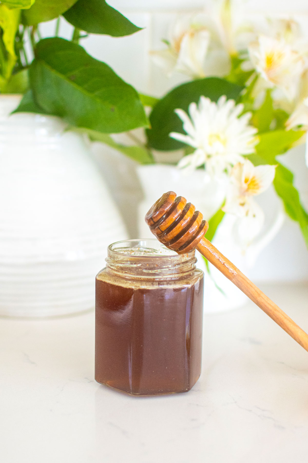 homemade organic hot honey in a glass honey jar with a honey dipstick on a marble counter with fresh flowers.
