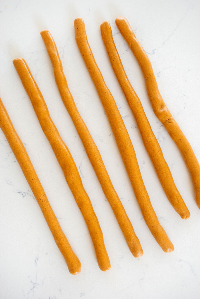 mini peanut butter oat dog treat dough formed into 6 ropes on a white countertop.