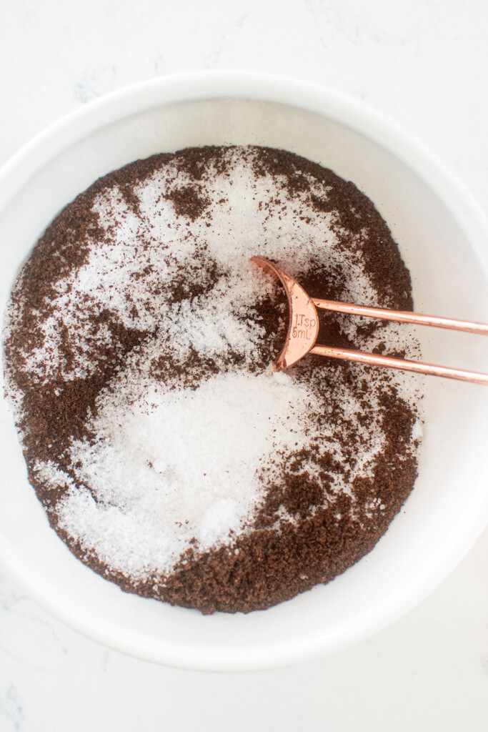salt and ground vanilla bean in a mixing bowl on a white marble counter being mixed with a copper measuring spoon.