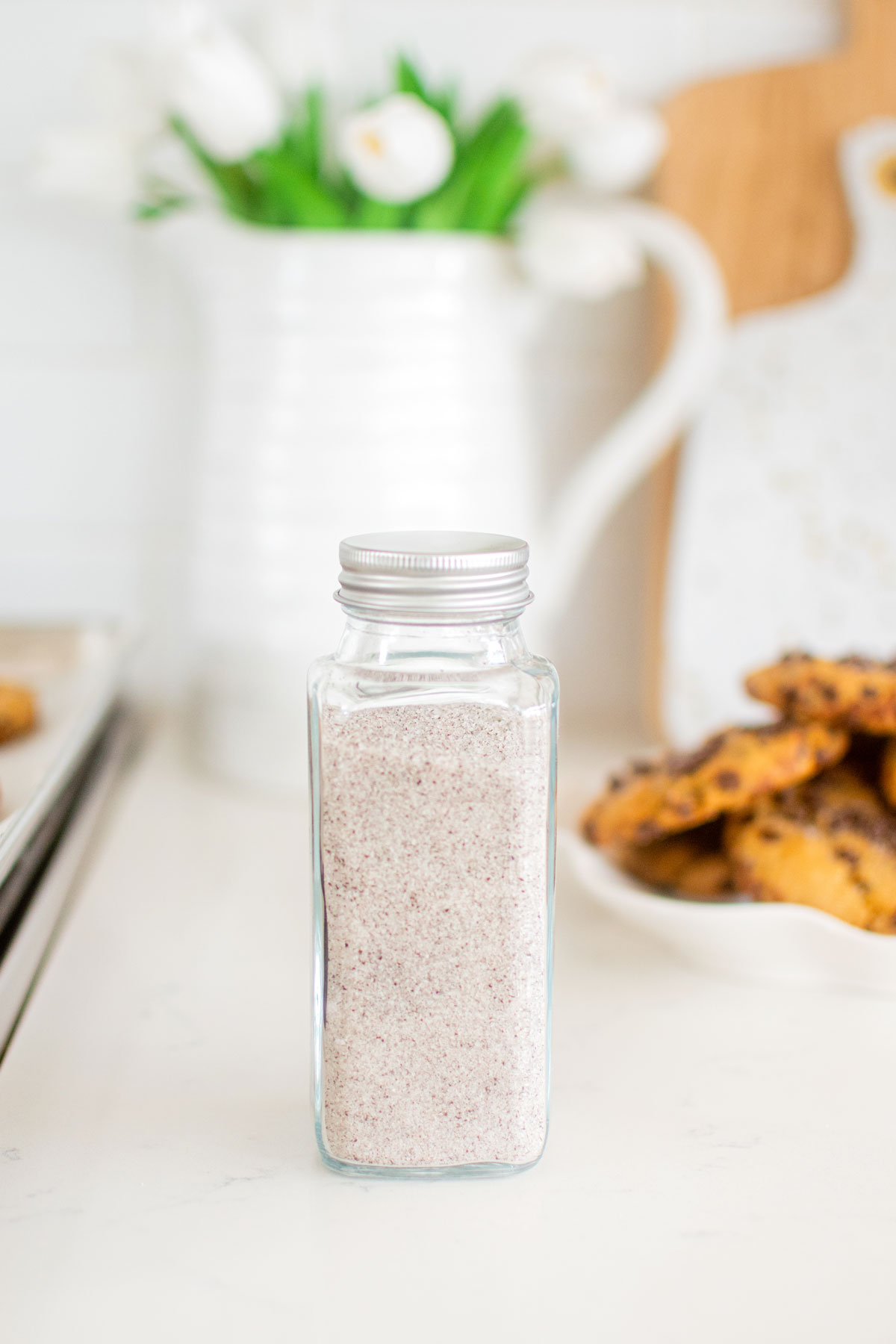 vanilla sea salt in a spice jar on a white marble counter with cookies and flowers in the background.