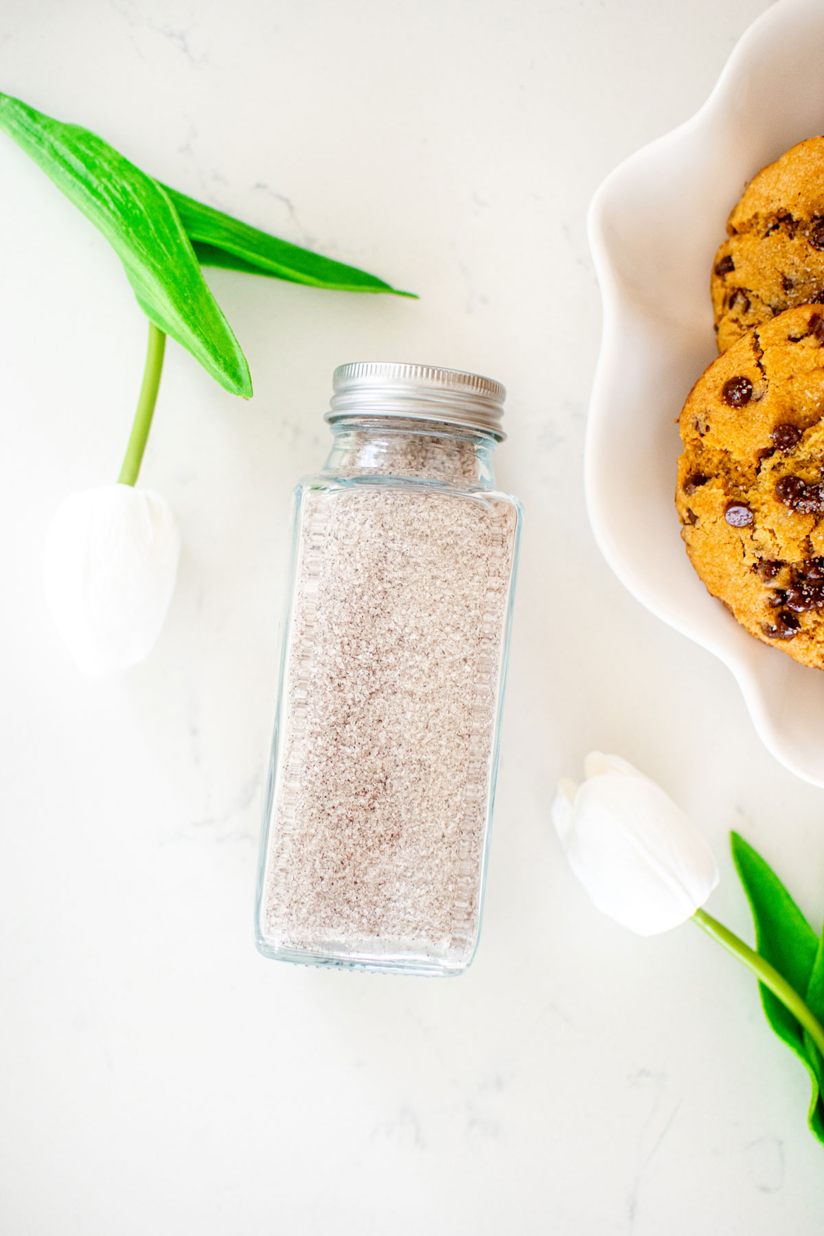 vanilla sea salt in a spice jar on a white marble counter with cookies and flowers in the background.