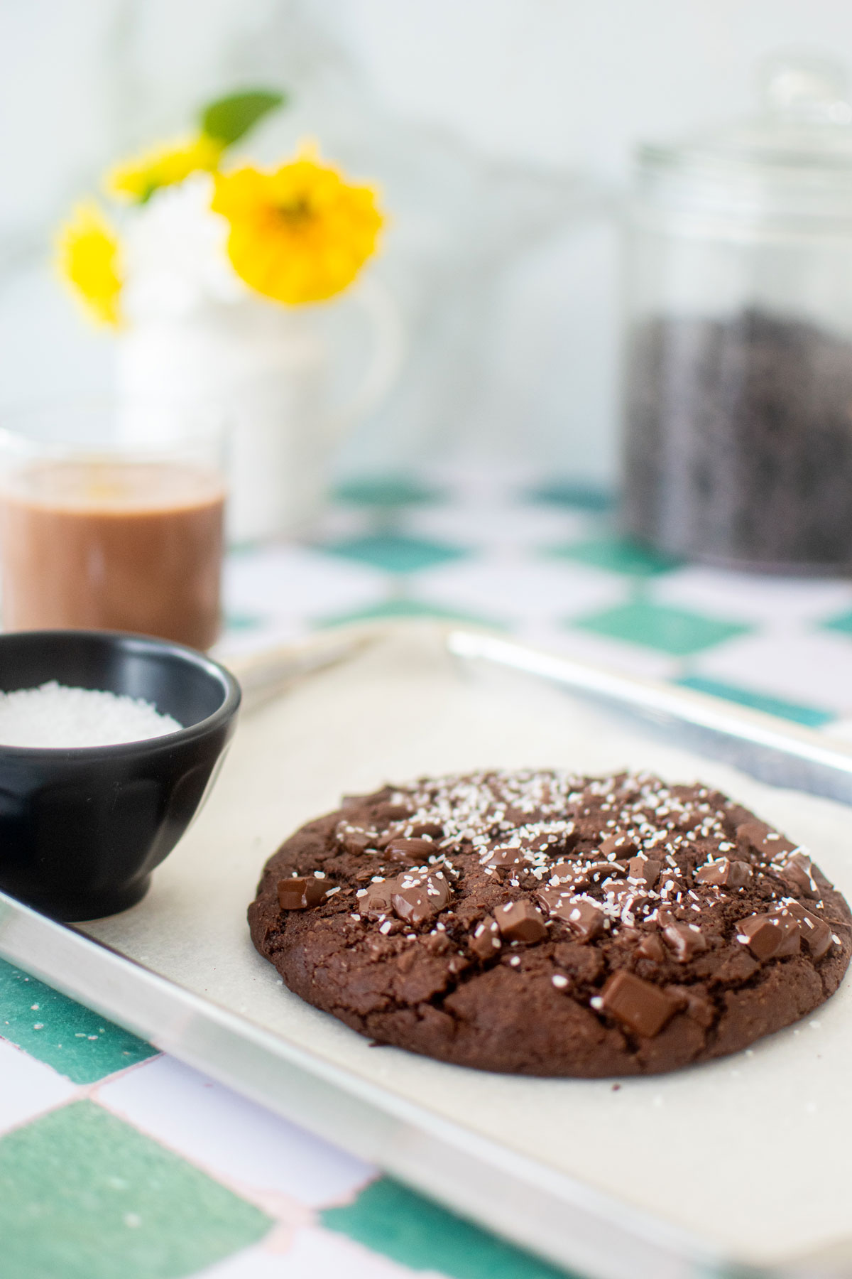 one big chocolate cookie on a baking sheet on a green tile counter with chocolate milk and fresh flowers.