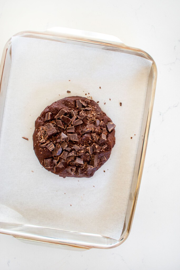 big chocolate cookie dough bursting with dark chocolate on a baking sheet on a white marble counter.