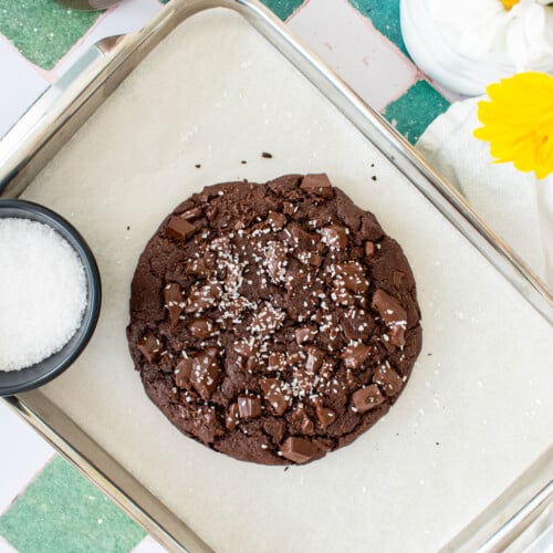 one big chocolate cookie on a baking sheet on a green tile counter with chocolate milk and fresh flowers.
