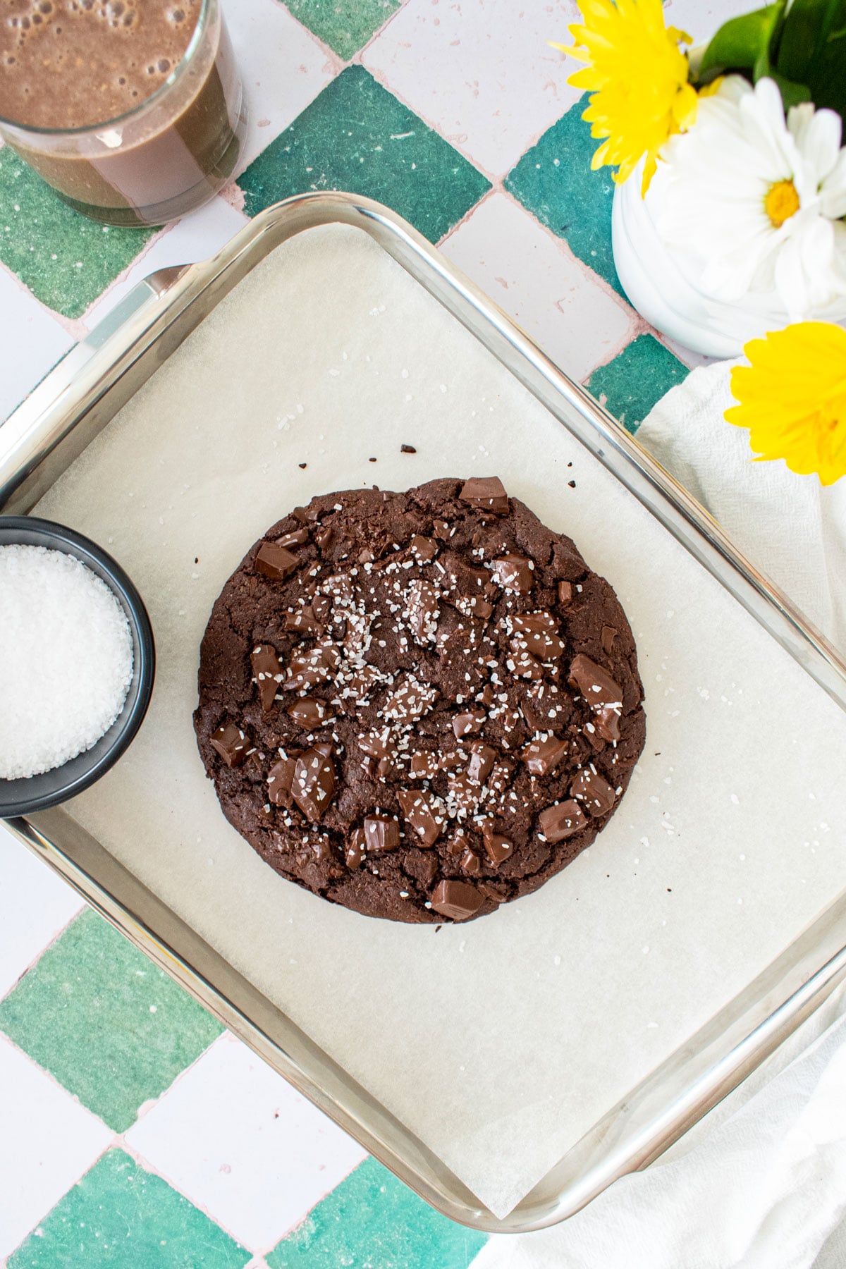 one big chocolate cookie on a baking sheet on a green tile counter with chocolate milk and fresh flowers.