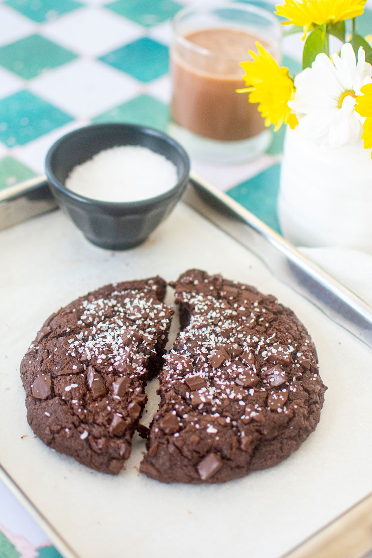one big chocolate cookie broke in half on a baking sheet on a green tile counter with chocolate milk and fresh flowers.