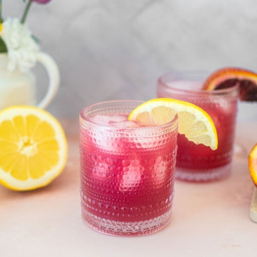 blood orange lemonade in a glass with a slice of lemon on the counter.