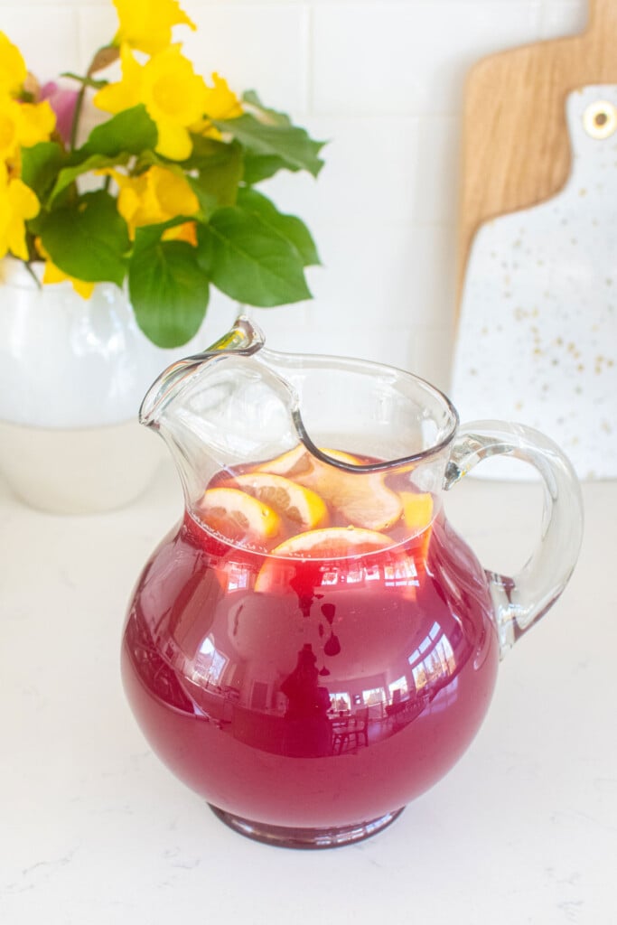 blood orange lemonade in a pitcher with lemon slices and a vase of flowers on a countertop.