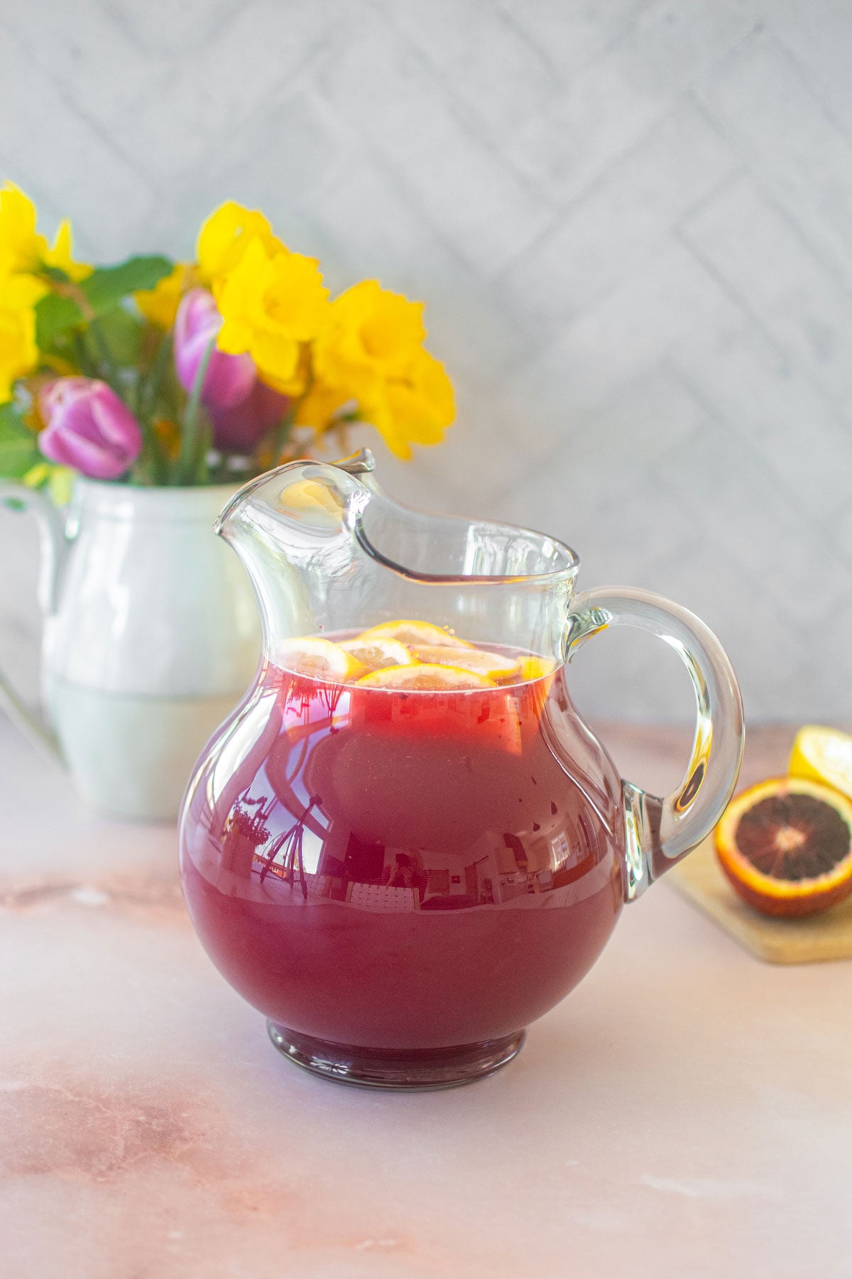 blood orange lemonade in a pitcher with sliced lemons on the counter with a vase of flowers.