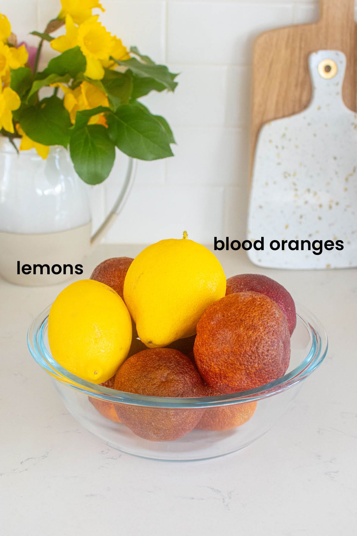 blood oranges and lemons in a bowl on the counter with a vase of flowers.