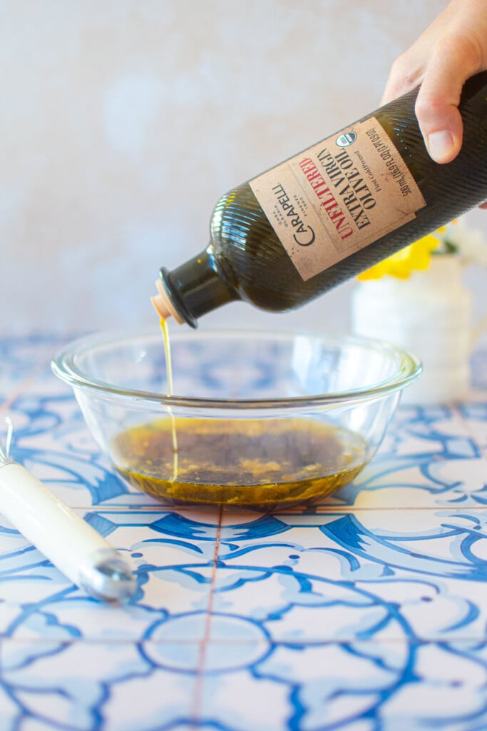 glass bowl with olive oil being poured into it on a blue tile counter.