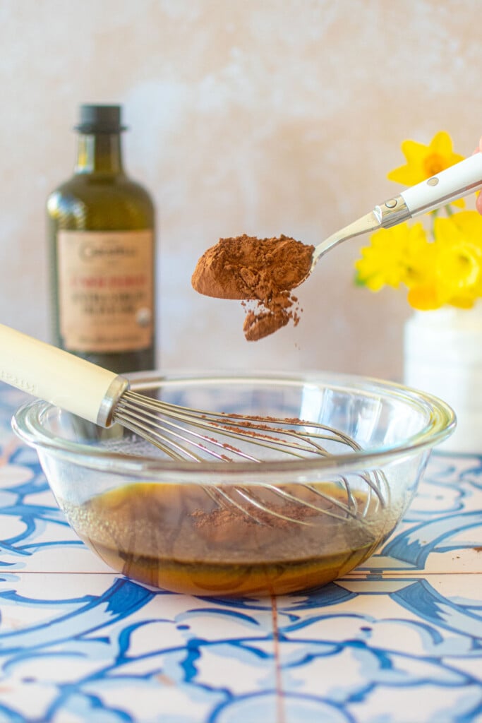 cocoa being added to a glass mixing bowl with olive oil on a blue tile table.