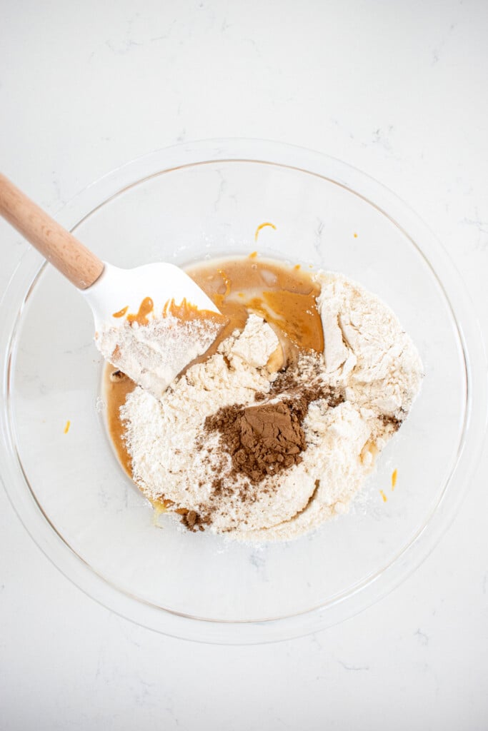 peanut butter, flour, coconut oil, honey and mushroom powder in a glass mixing bowl on a white counter.