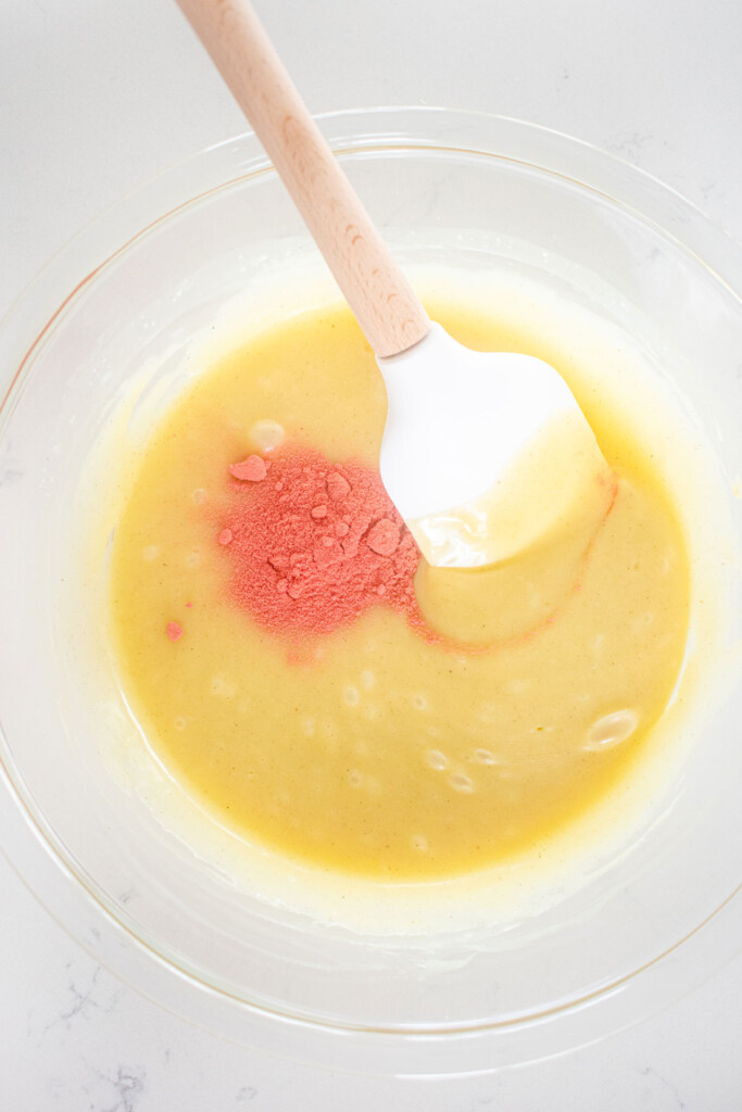white chocolate ganache with pink strawberry powder in a glass mixing bowl with a spatula on a marble counter.