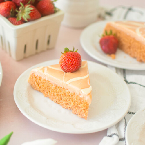 strawberry snack cake with pink frosting and a strawberry on top on a plate on a pink table with a berry basket of strawberries and fresh flowers.