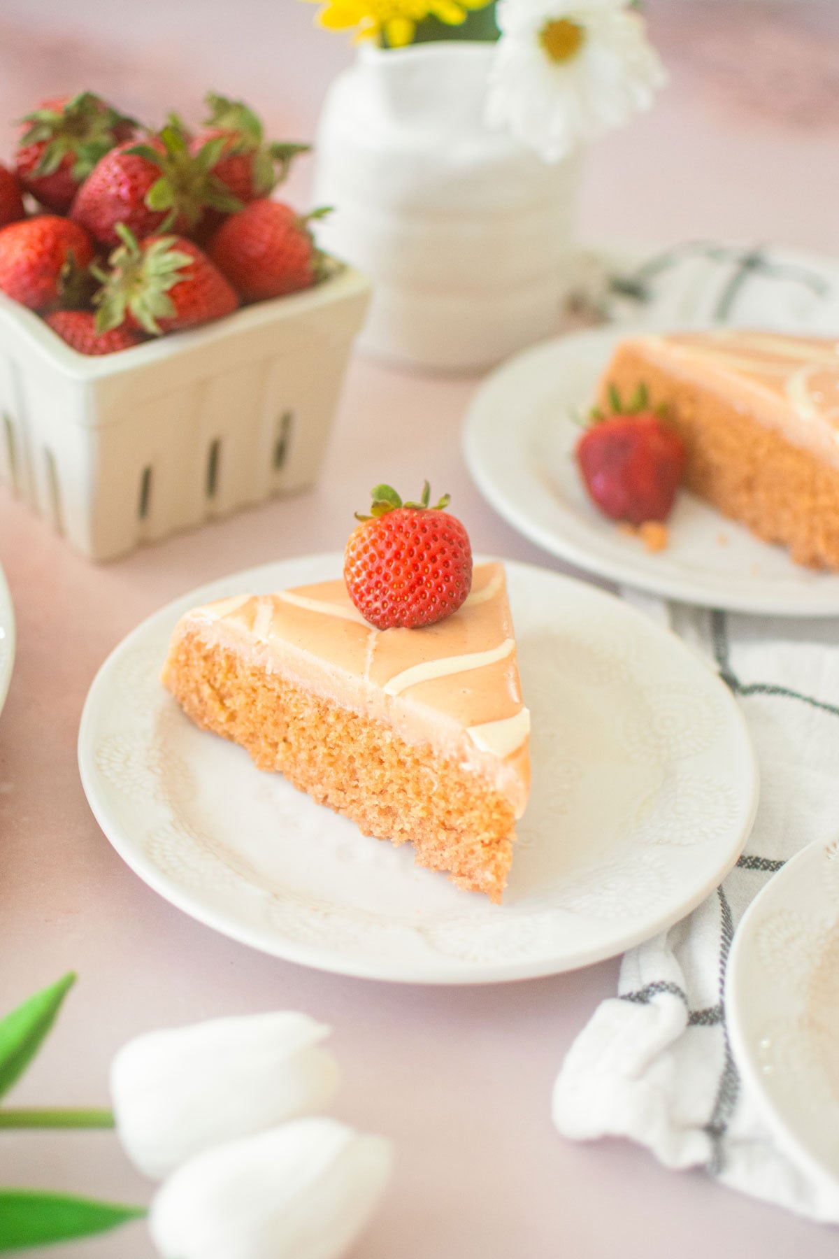 strawberry snack cake with pink frosting and a strawberry on top on a plate on a pink table with a berry basket of strawberries and fresh flowers.