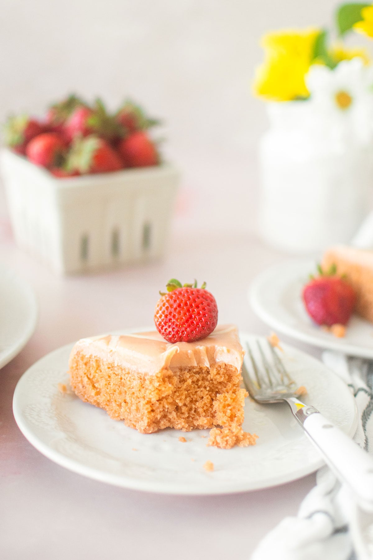 strawberry snack cake slice on a plate with a bite taken out on a pink table with fresh strawberries and flowers.