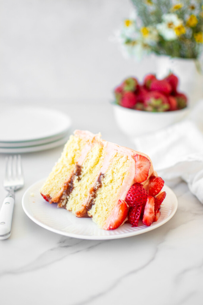 slice of strawberry vanilla layer cake on a white cake plate on a marble table with fresh flowers.