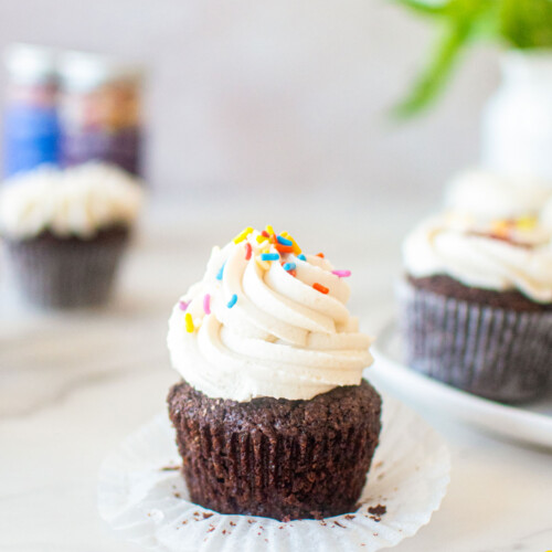 vegan chocolate cupcake with vanilla frosting and rainbow sprinkles on a white marble counter with fresh flowers.