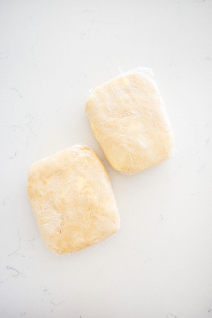 two rectangle pie doughs wrapped in plastic wrap on a white marble countertop.