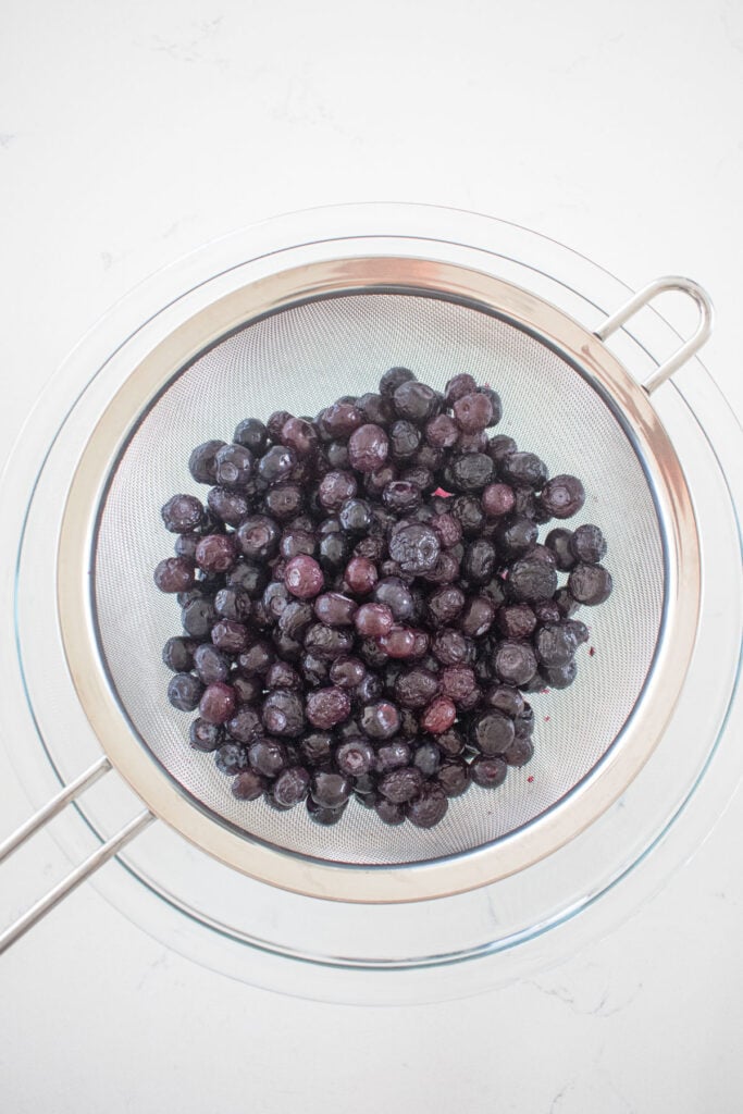 blueberries straining in a fine mesh strainer in a glass mixing bowl on a white marble countertop.
