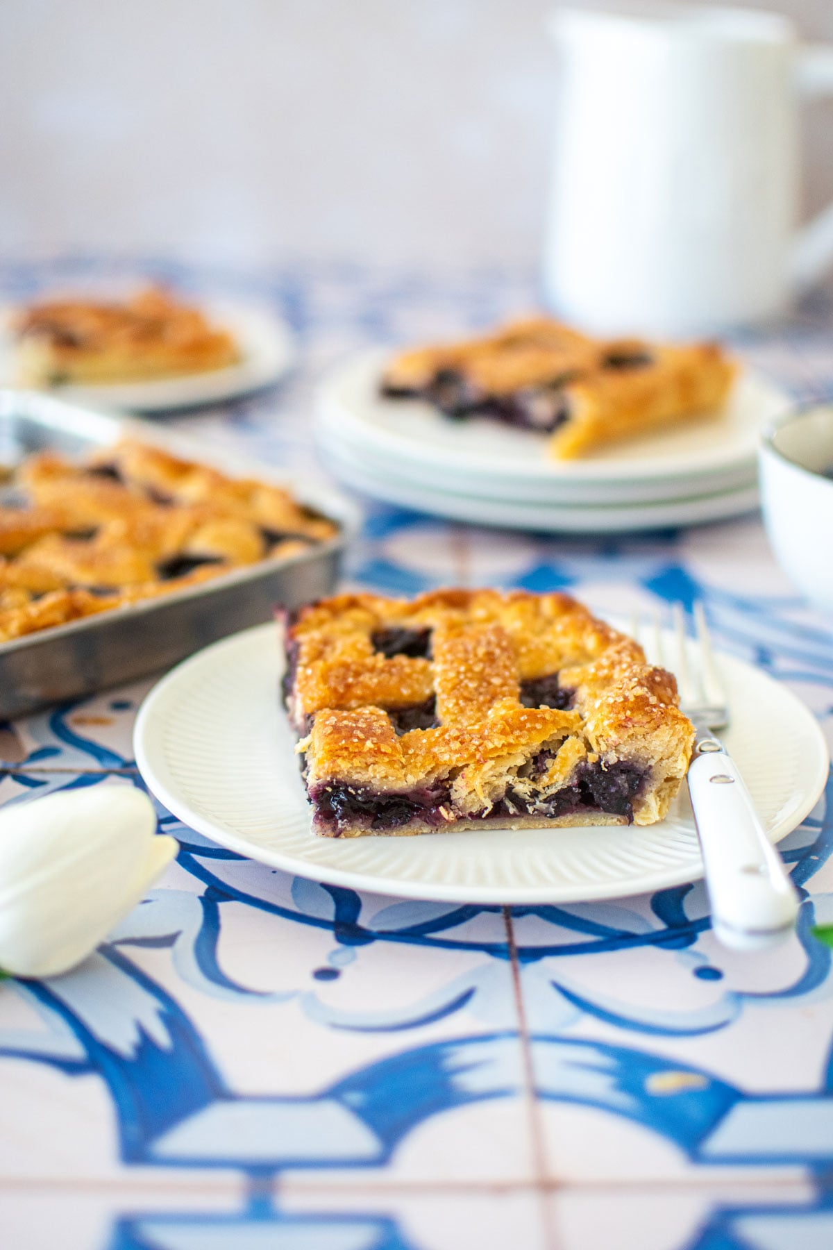 blueberry slab pie without eggs on a white plate with a fork on a blue tile countertop.