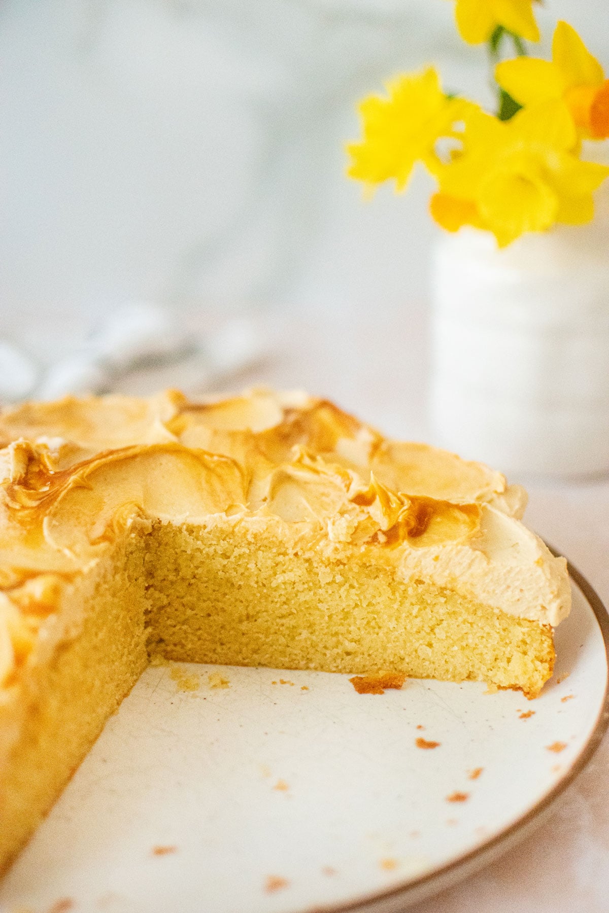 caramel buttercream frosting on vanilla cake sliced on a cake plate on a tan counter with fresh flowers.