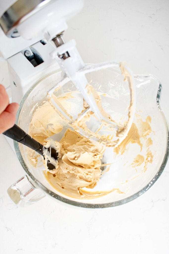 caramel buttercream frosting being stirred in a glass stand mixing bowl on a white marble counter.