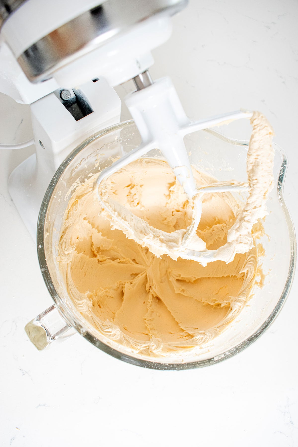 creamy caramel buttercream frosting in a glass stand mixing bowl on a white marble counter.