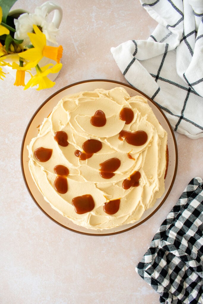caramel buttercream frosting with dollops of caramel sauce on a single layer cake on a plate on a tan counter with fresh flowers.