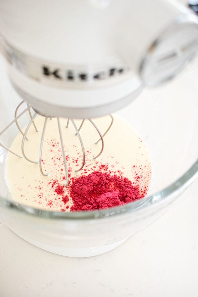 heavy whipping cream and freeze dried raspberry powder in a glass stand mixing bowl on a white marble counter.